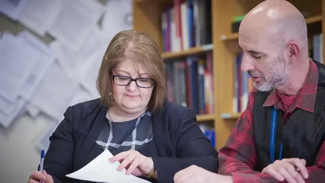 Two professionals review documents together in a research or academic setting, surrounded by bookshelves and pinned papers on the wall.