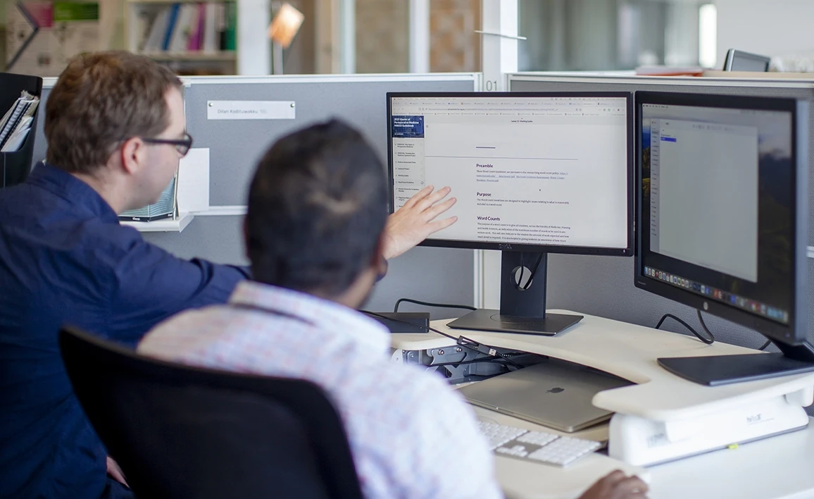 Two professionals collaborate at a workstation, discussing publications displayed on a computer screen in a modern office setting.