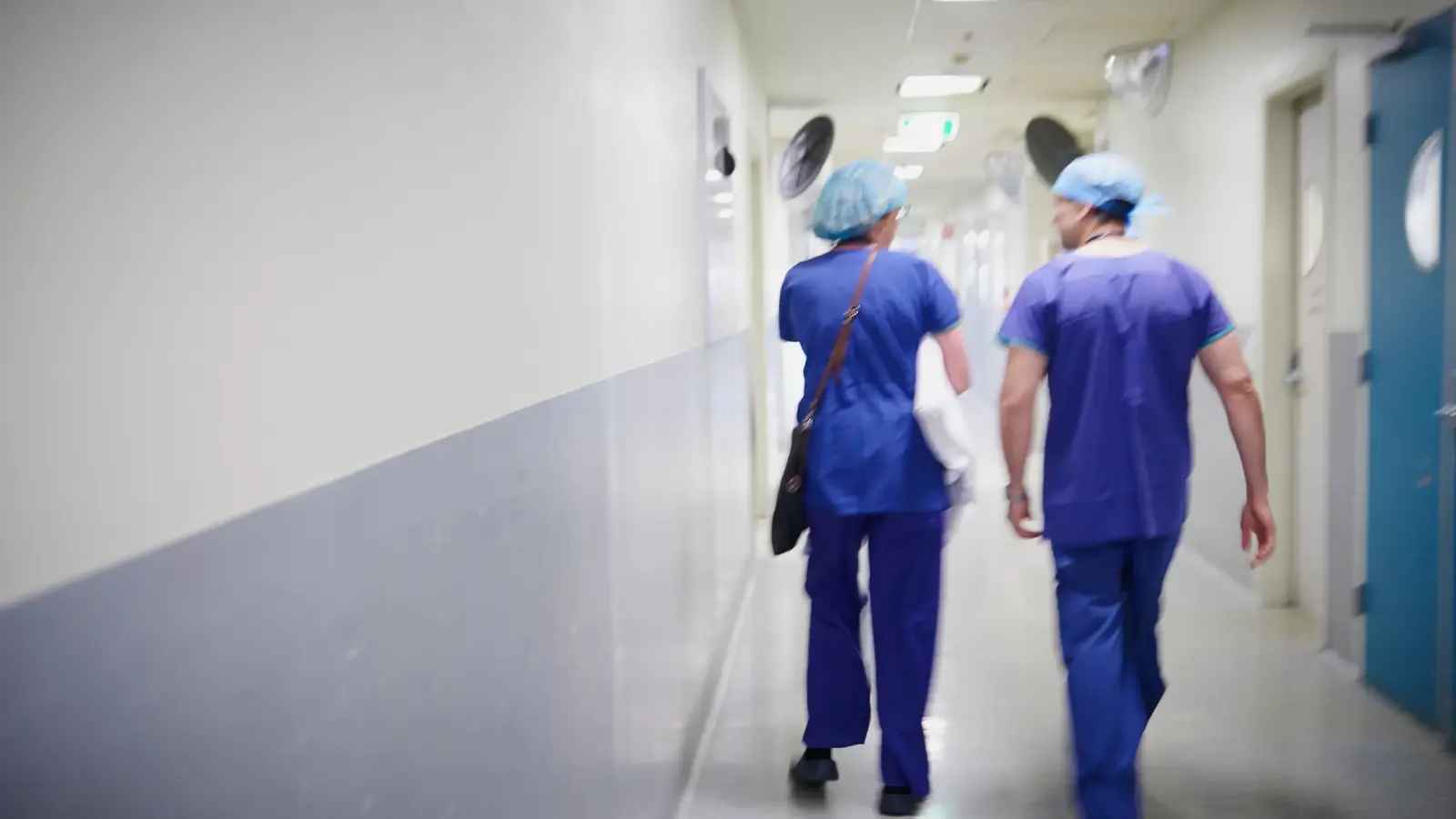 a male and female doctor in scrubs walk down a hospital corridor, away from the camera