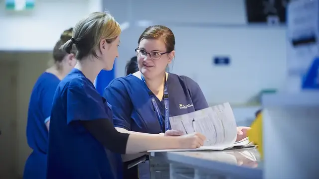 Two healthcare professionals in navy scrubs discuss patient documentation at a hospital workstation, with a busy clinical environment in the background.