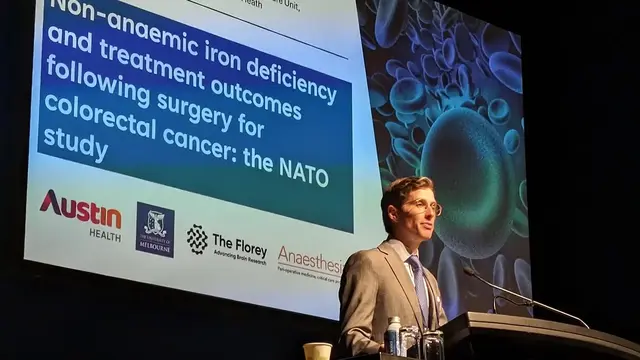 An anaesthetist in a suit stands at a podium, delivering a talk on the NATO study about non-anaemic iron deficiency and treatment outcomes after colorectal cancer surgery, with a large presentation screen behind him.