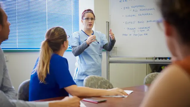 Person in scrubs in front of whiteboard talking to students sitting around a table