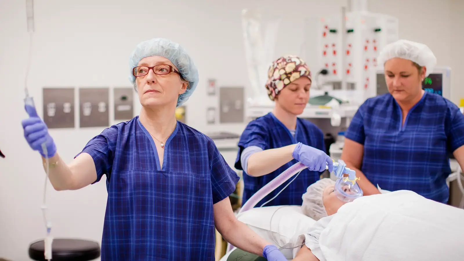 Person in scrubs checking IV fluids. Two people in scrubs in the background check on patient with anaesthesia mask on