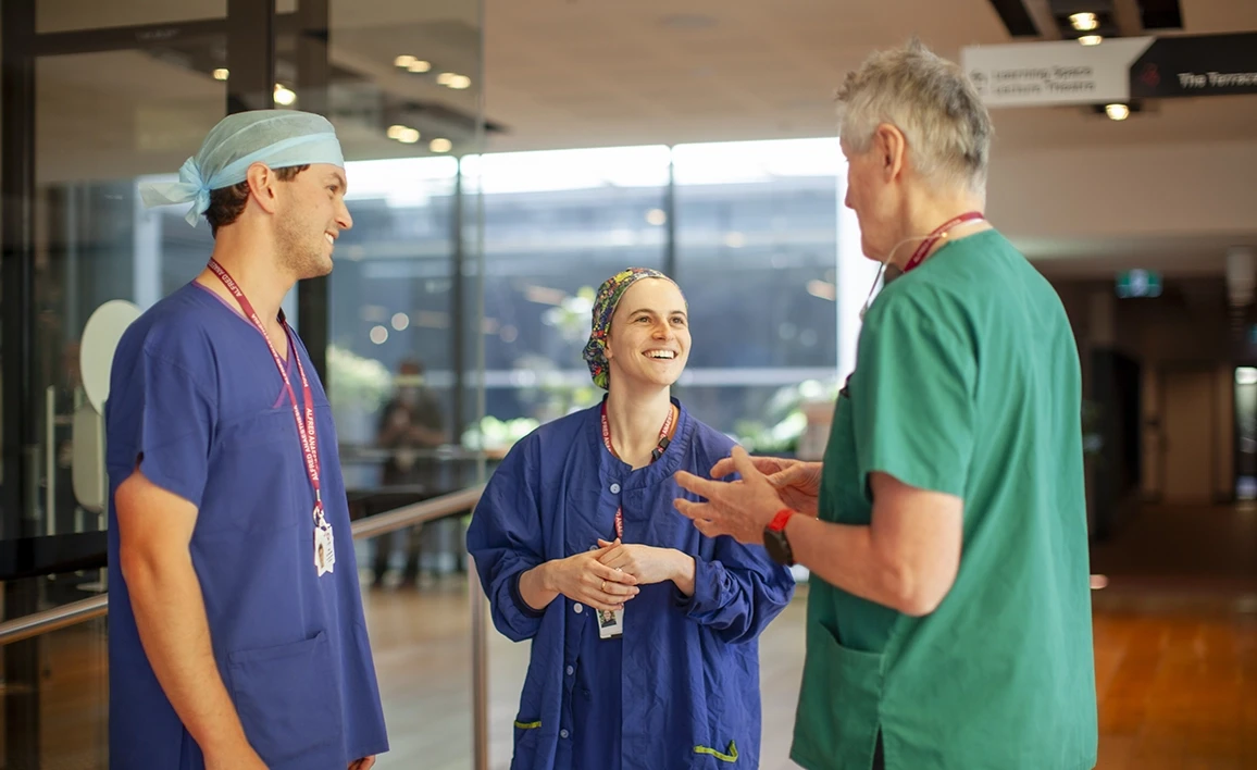 The image shows three healthcare professionals in scrubs having a lively and friendly conversation in a hospital or clinical setting.