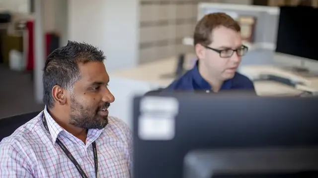 The image shows two men working side by side in an office setting, focused on computer screens. One is wearing a checkered shirt and speaking, while the other, in a dark shirt and glasses, is concentrating on his screen.