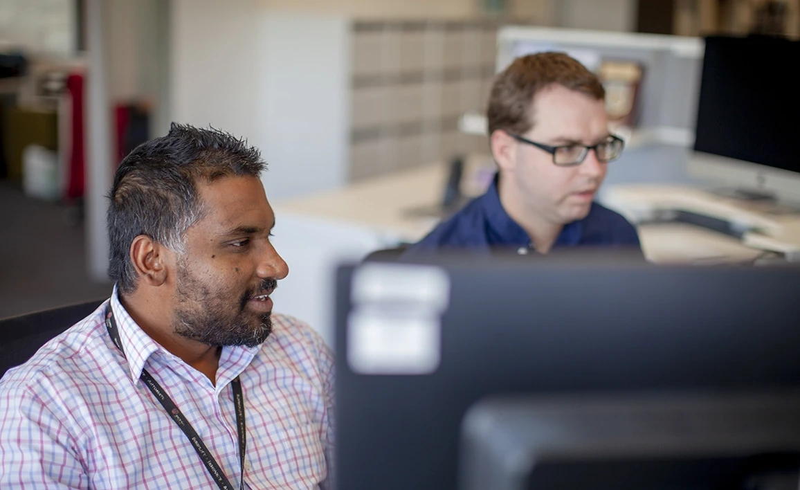 The image shows two men working side by side in an office setting, focused on computer screens. One is wearing a checkered shirt and speaking, while the other, in a dark shirt and glasses, is concentrating on his screen.