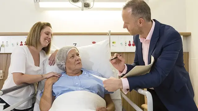 A patient in a hospital bed engaging in discussion with an anaesthetist in a suit, while a supportive family member sits beside them, smiling.