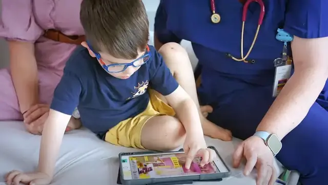 A young child wearing glasses interacts with a tablet while sitting on a hospital bed, accompanied by a caregiver and an anaesthetist in scrubs with a stethoscope.