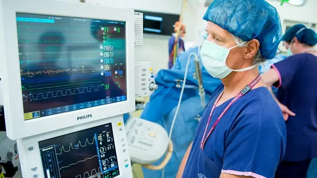 A masked and capped anaesthetist monitors vital signs on a screen in a busy operating room, with surgical staff working in the background.