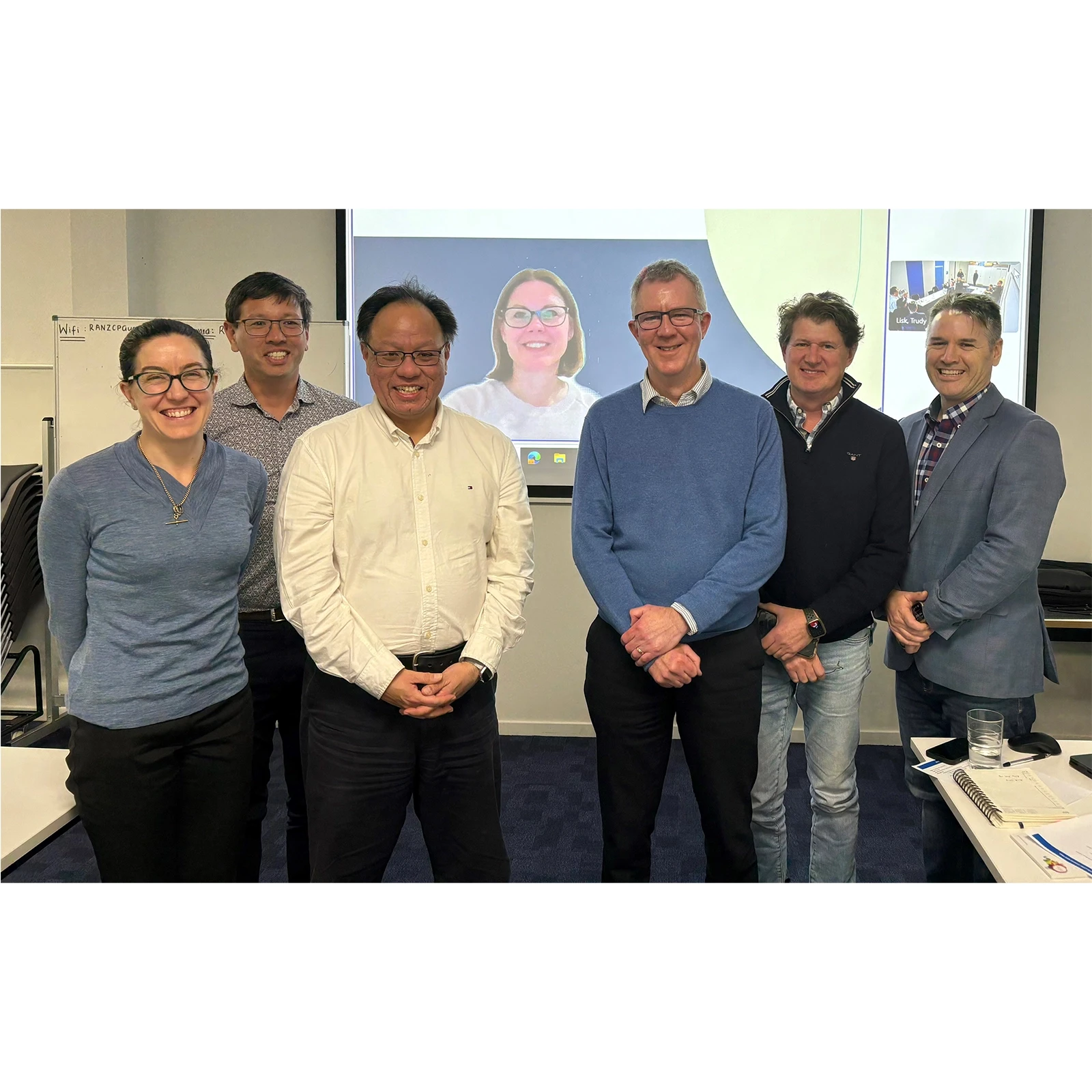 Six people standing and one on a screen behind