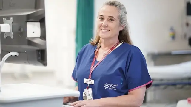 A smiling research coordinator in scrubs stands at a workstation in a hospital setting, ready to assist with patient care.