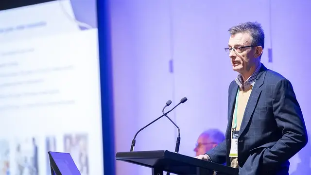 A professional speaker at a conference, presenting at a podium with a large screen displaying slides in the background.