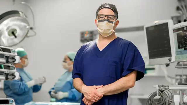 Male anaesthetist facing camera in operating theatre, wearing scrubs. Other doctor's chat in the background. 