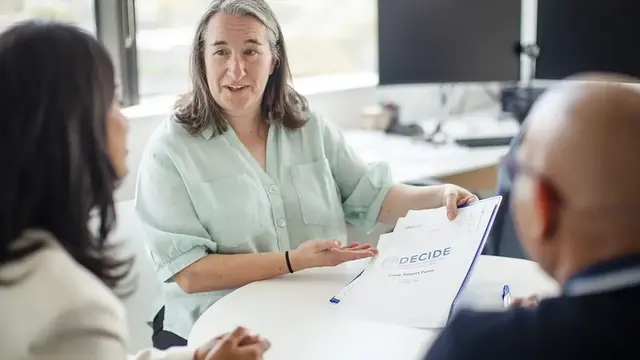 A research coordinator in a light green blouse discusses a document labeled "DECIDE Case Report Form" with a father and daughter in a modern office setting.