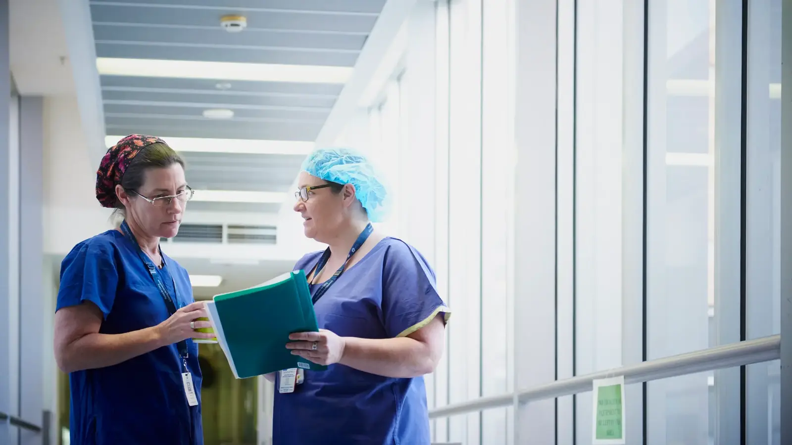 Two people in blue scrubs talking in a hospital corridor. One is holding a folder of papers