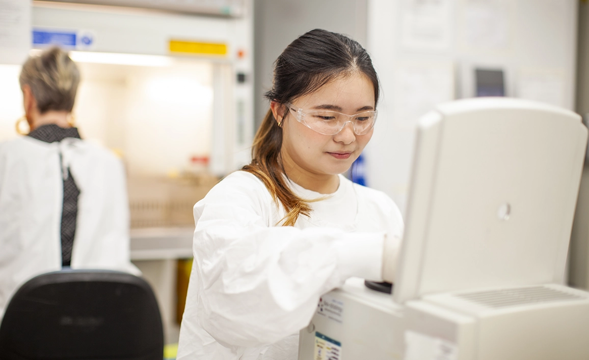 A scientist in a lab coat, safety glasses, and gloves operates laboratory equipment, while another person works near a fume hood in a research lab.
