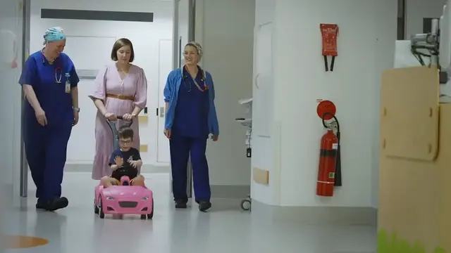 A young child rides a pink toy car down a hospital hallway, accompanied by a caregiver and two anaesthetists in scrubs, creating a supportive and comforting environment.