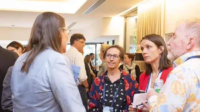 A professional networking event where four individuals engage in conversation, with other attendees mingling in the background.