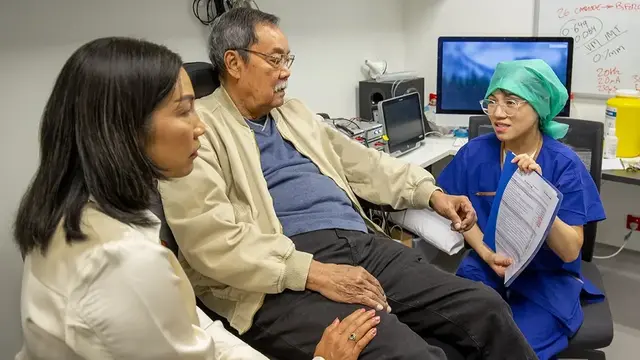 A research coordinator in blue scrubs discusses a medical document with a patient and a companion in a clinical setting, surrounded by medical equipment.