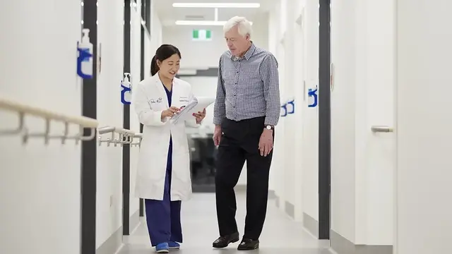 An anaesthetist in a white coat walks alongside an elderly patient in a medical facility, discussing paperwork in a bright, modern hallway.