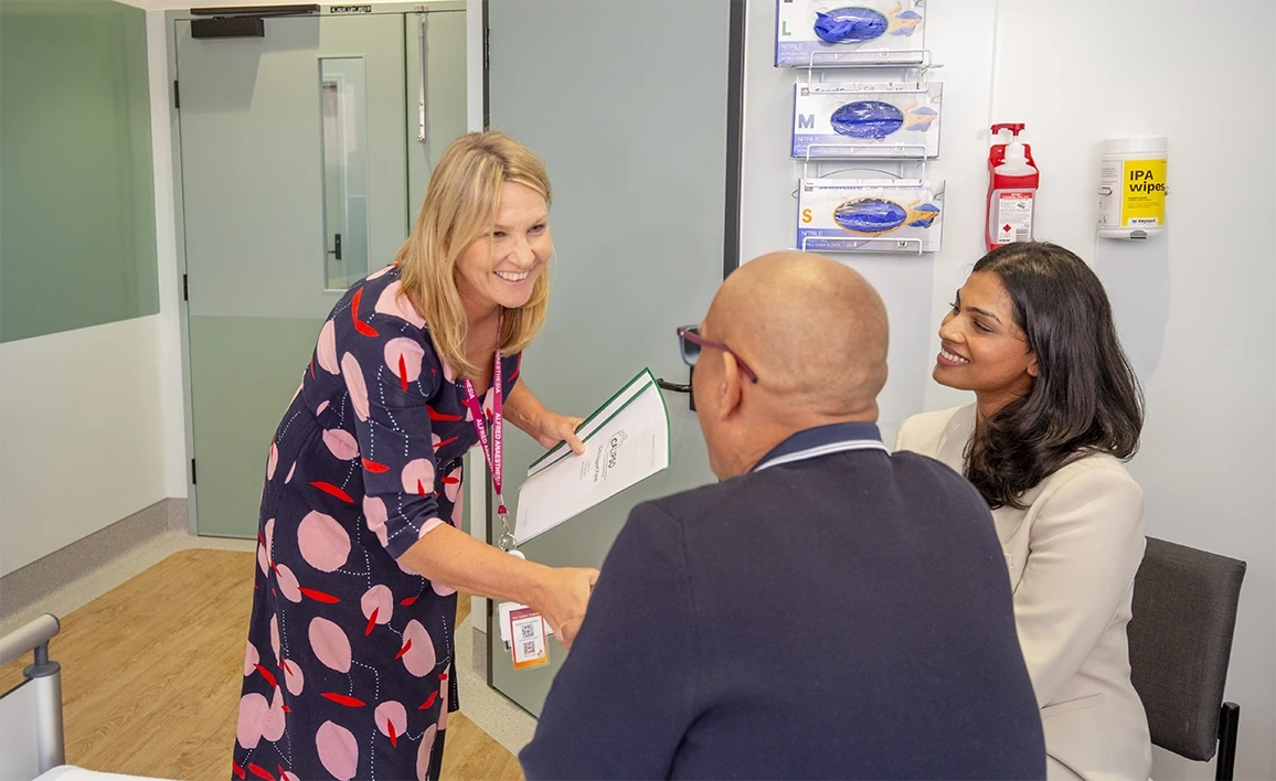 The image shows a friendly interaction in hospital setting. A staff member is warmly greeting a seated man with a handshake, while another family member looks on smiling.