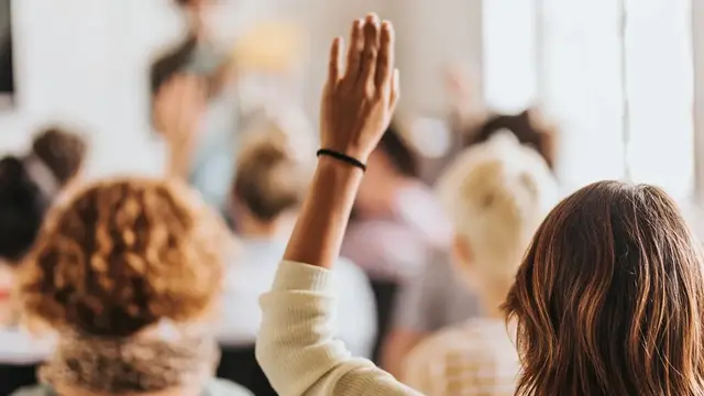 Woman raising hand in a crowd, facing a speaker