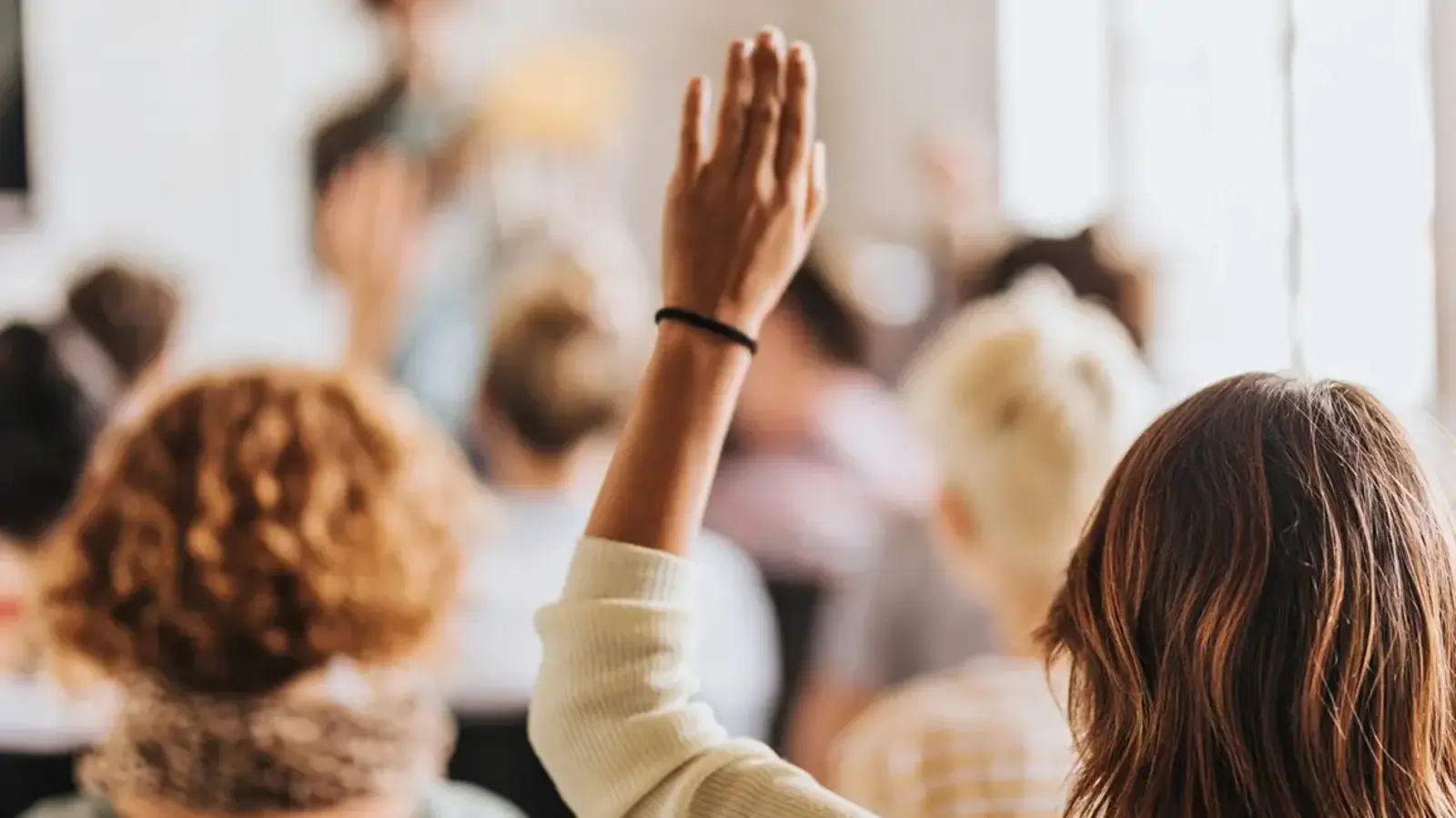 Woman raising hand in a crowd, facing a speaker